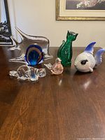 Six animal figurines arranged on wooden table: clear glass dog lying down, translucent blue and amber fish with clear wings, iridescent pink bird, green opaque cat, blue and white ceramic fish with black eyes, green glass cat with surface pock marks.