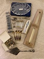Photo shows all items in the lot arranged on fabric: Mikasa crystal plate, white taper candles boxed, hand-blown glass handled cheese cutter, metal apple place card holders, silver plate serving utensils, small decorated spoons and others.