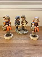 Front view of all three Hummel figurines displayed on wooden surface showing two girls with baskets and the boy and girl couple figurine on a base.