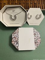 Photo shows full necklace with cubic zirconia stones in a semicircular pendant, rhodium plated silver tone metal, along with matching earrings in original packaging.