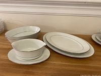 Photo shows two open vegetable bowls, two oval platters, and a gravy boat with attached underplate, all white porcelain with delicate raised floral pattern and silver trim.
