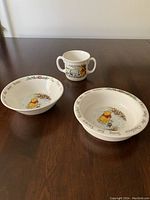 Photo showing full set of two bowls and one two-handle mug on wooden table, displaying Winnie the Pooh designs and text.