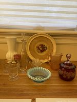 Full view of the vintage glass items arranged on wooden surface in front of window. Shows white vase, goblets, decorative plate, peach pitcher, clear dishes, blue ruffled bowl, and ruby red covered jar.