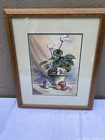 Framed watercolour painting showing a cyclamen plant with leaves and flowers, displayed against a wall.