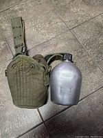 Image of the aluminum military canteen next to its green canvas cover and attached belt laid flat on the floor.