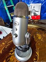 Blue Yeti USB Microphone on a decorative wooden table with a blue tarp background, showing front and side views, displaying mute and volume controls, USB cable wrapped around stand base.