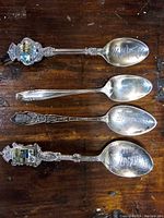 Four sterling silver spoons arranged side by side on wood surface showing front details including enamel crests and engravings on bowls.