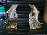 Frontal view of two brass dolphin bookends showing dolphins rising from stylized ocean waves, placed side by side.