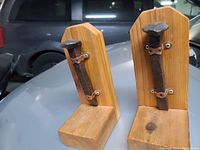 Pair of two wooden bookends with large rustic iron nails affixed to each wooden piece using copper brackets, placed on metallic surface near vehicle.
