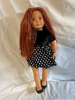 A doll with long auburn hair, wearing a black velvet top and black skirt with white polka dots and black shoes.