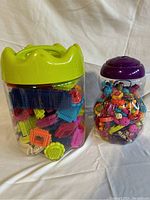 Two clear jars containing plastic building kit pieces. One jar has a green lid and contains multicolored bristle-style pieces. The other jar has a purple lid and contains various colorful beads and flower-shaped components for jewelry making.