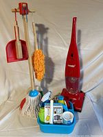 Full view of wooden broom seat on stand with mop, dustpan, brush, plastic play cleaning supplies caddy, and Eureka plastic kids vacuum.