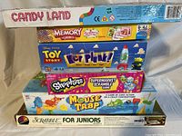 Photo of seven boxed children's board games stacked for sale including Candy Land, Memory game, Toy Story Ker Plunk, Shopkins Supermarket Scramble, Mouse Trap, and Scrabble for Juniors.