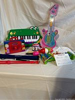 Photo of the full lot showing green frog keyboard, pink Leap Frog Touch Magic guitar, red slide whistle, red music box, maracas, and other accessories on a light fabric background.