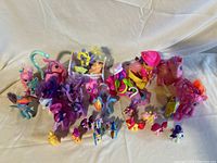 Group of My Little Pony dolls and figures arranged on a white cloth background showing a variety of sizes, colors, and types including larger plush ponies and smaller figurines.