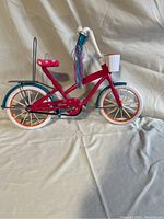 Photo of a red metal frame doll bike with teal and orange wheel details, pink seat, white basket, and multi-color handlebar streamers against a white fabric background.