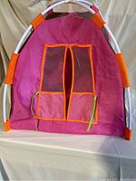 Front view of the pink and orange doll tent with window flaps and white curved poles visible.