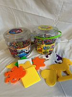 Two clear plastic containers filled with colorful beads and assorted shaped plastic pegboard templates arranged on a white cloth surface.