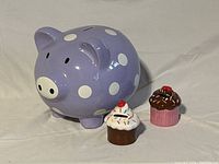 Photo of one large purple piggy bank with white polka dots alongside two smaller cupcake-shaped piggy banks, one with white frosting and sprinkles, the other with chocolate frosting.