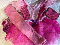 Pink and purple child's dress with sequin ornaments and tutu skirt, pink sequin purse, long pink scarf, and pink jewelry including necklace and bracelets arranged on flat surface.