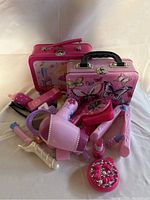 Pretend hair and makeup items in pink and purple including hairdryer, hair straightener, lipstick, and decorative metal storage cases with butterfly and cartoon designs.