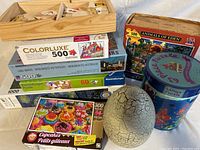 Photo showing stacked boxed cardboard puzzles and a wooden box with large wooden puzzle pieces with painted animal faces, plus a decorative egg