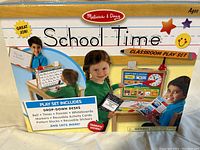 Box showing Melissa & Doug School Time Classroom Play Set with children playing and listing included items such as drop-down desks, bell, timer, pointer, whiteboards, markers, reusable activity boards, pattern blocks, and extras like magnetic pattern blocks kit.