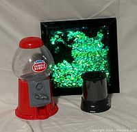 Photo of three items: red Dubble Bubble gum ball machine, black night light star projector, and green sequin wall art in black frame against a cloth background.
