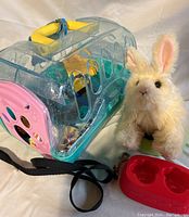 Photo showing clear plastic pet carrier with yellow handle and black strap, soft Fur Real electronic bunny toy, and red pet food bowl.