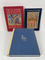 Three hardcover facsimile books showing illuminated manuscript art, including a red slipcover and blue hardcover volume