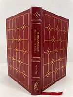 Front and back cover of the book showing deep red leather binding, gold gilt decoration, spine with title and author name embossed in gold, and a silk bookmark.