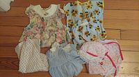Five assorted baby doll dresses shown laid out on a wooden floor including floral, sunflower print, pastel, and patterned designs.