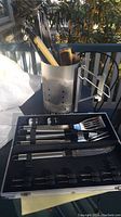 View of the barbecue tools including charcoal chimney starter with wood handle utensils inside, and metal BBQ tool case open showing stainless steel utensils organized inside.