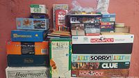 Full view of stacked assorted board games showing major titles including Monopoly, Trivial Pursuit, Carcassonne, HuggerMugger, Clue, Sorry, and various card and puzzle games.