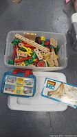 Plastic container filled with wooden Brio Mec construction parts, including beams, wheels, screws, connectors in various colors, accompanied by a Brio Mec instruction booklet and vehicle kit carrying case.