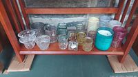Photo showing a variety of candle holders and glass containers in clear, amber, pink, teal, cream and dark colors organized on a wooden shelf.