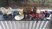 Wide shot of all ceramic pottery items displayed on porch railing including bowls, mugs, vases, and decorative pieces.