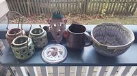 Full view of all ceramic and pottery items on a table including the candle holders, face vase, pitcher, bowl, and lid.