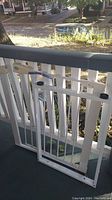 White metal child safety gate leaning against white railing outdoors, showing vertical bars, mounting hardware, and plastic end caps.