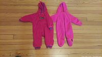 Photo shows two fleece children's snowsuit onesies for winter, one bright pink and one darker pink with blue lining and patterned cuffs/feet.