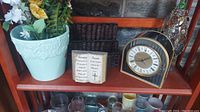 Overview of shelf with ceramic flower pot, religious ceramic plaque, wicker holder, vintage-style clock and decorative bottle