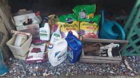 Photo showing various gardening supplies including bags of potting mix, bottles of garden sprays, a blue watering can, garden tools in a wooden box and additional items on gravel ground.