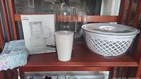 Photo showing all items included in the lot: glass carafe with box, plastic pitcher, dish towels, tea steeper, and salad bowl on wooden shelf.