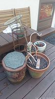 Photo showing large ceramic planter with sun relief design, terracotta planter with soil containing metal garden stakes, metal plant stands with glass bowls, and white ceramic planter.