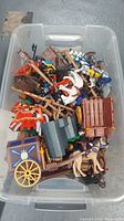 Clear plastic bin filled with assorted Playmobil medieval figures and accessories including horses, wagons, and weapons.