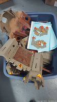 Photo shows a large blue plastic bin containing multiple large plastic Playmobil castle wall and tower pieces in brown and beige with flame decals, along with printed instruction sheets depicting assembly instructions.