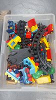 Clear overhead view of various colorful Playmobil train pieces including locomotives, cars, tracks, and accessories in storage bin.