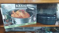 Photo showing two roaster pans side by side, one boxed Basic Essentials oval roaster with rack inside and one blue speckled rectangular roaster pan with lid