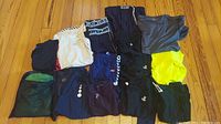 All athletic tops and shorts laid flat on wooden floor showing variety of colors and brands including Under Armour