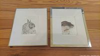 Two framed animal artwork prints side by side featuring an owl and an otter on a wood floor.
